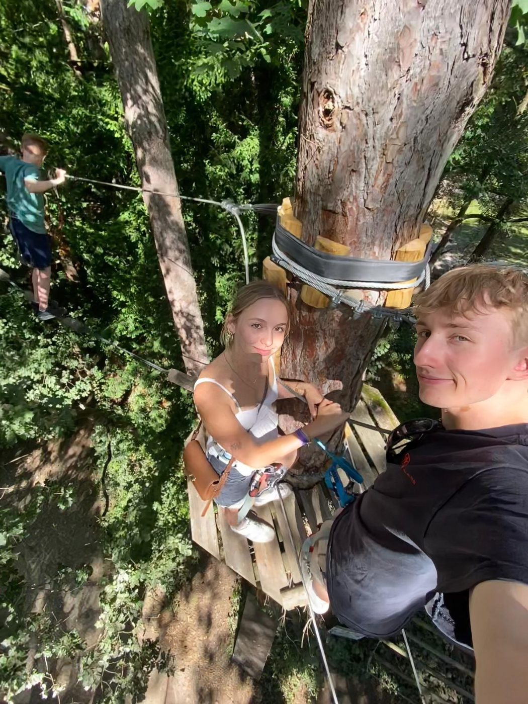 Selfie en hauteur dans un parcours accrobranche, elle sur une plateforme autour d'un tronc.