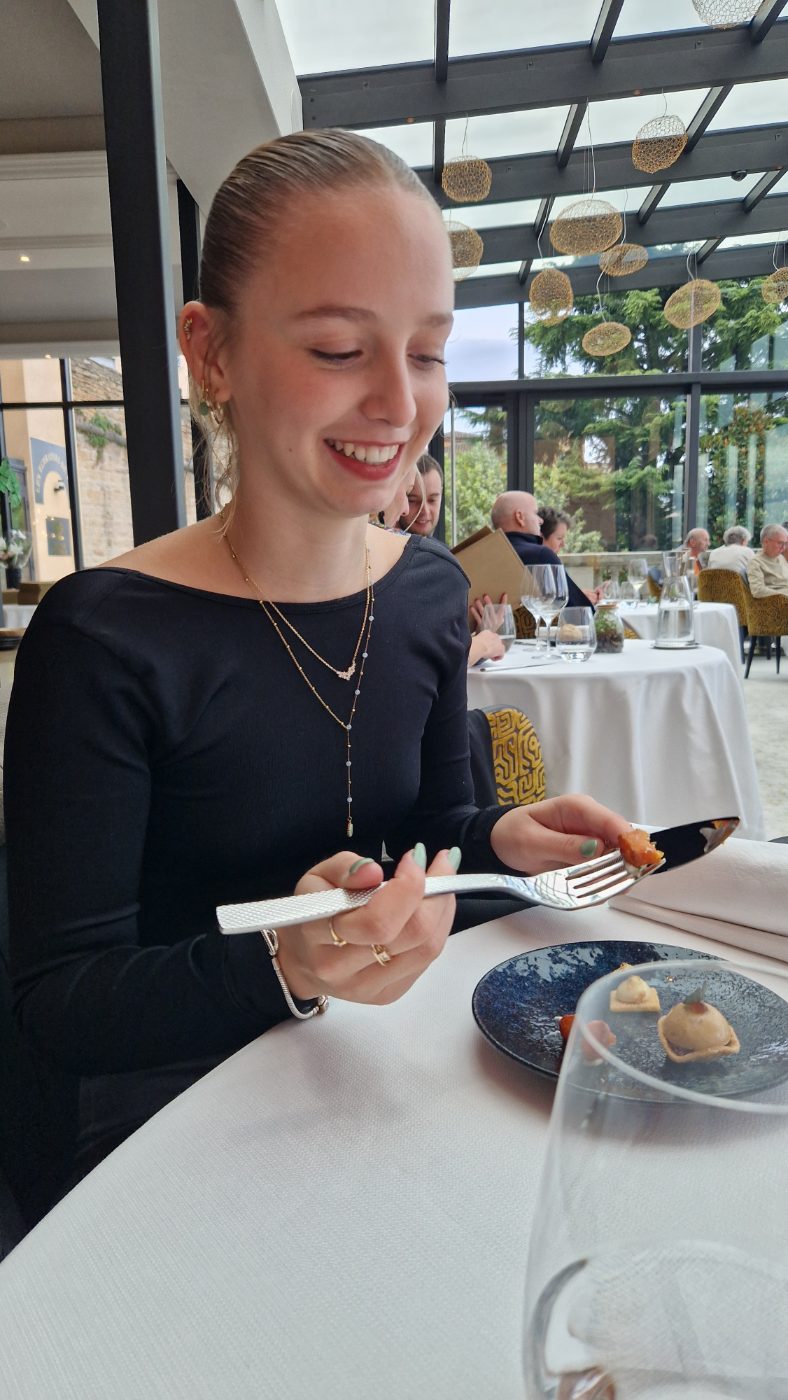 Elle rit devant une assiette dans une salle de restaurant sous verrière, nappe blanche.
