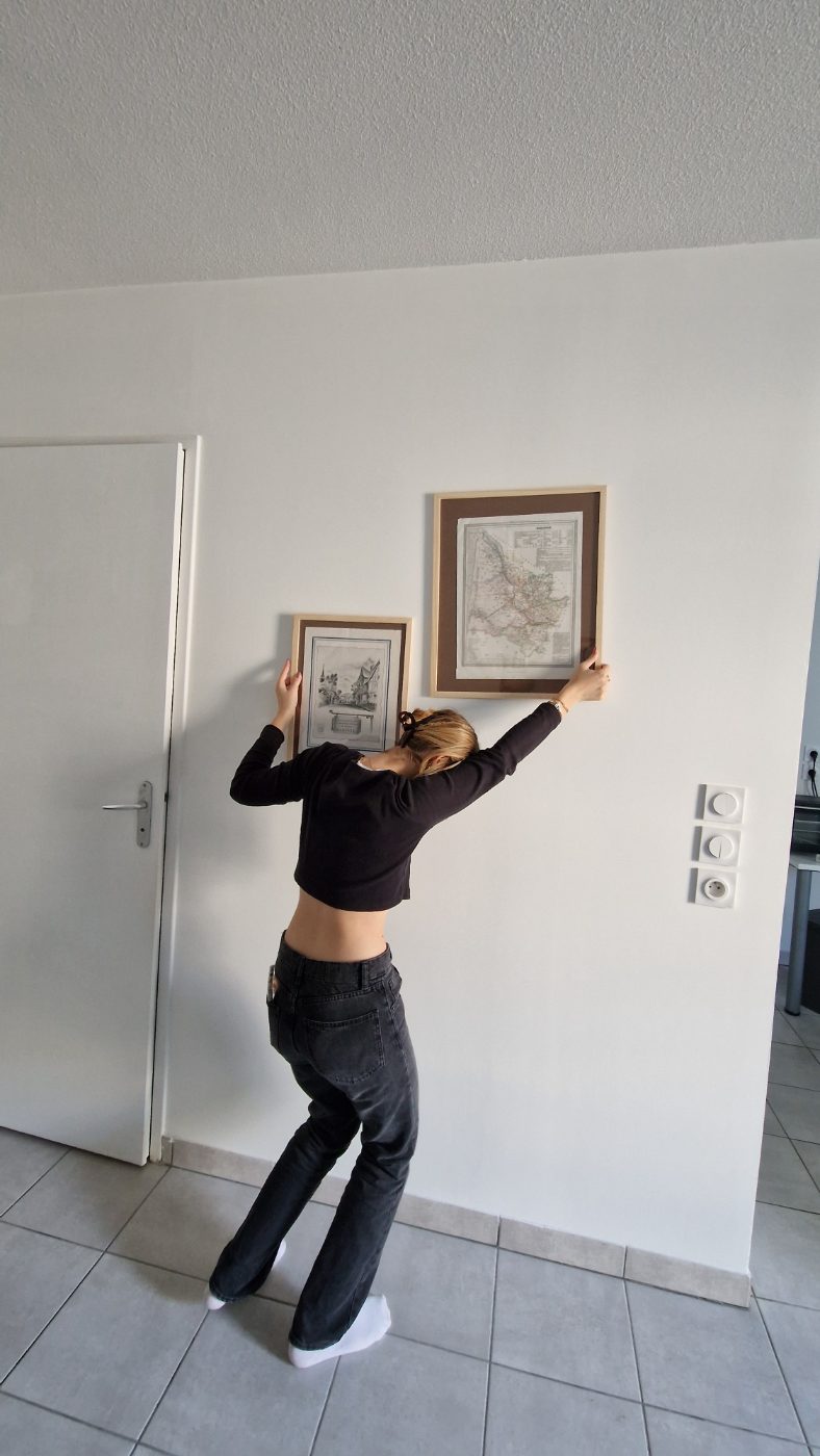 Vue de dos : elle tient à bout de bras deux cadres contre un mur blanc vide.