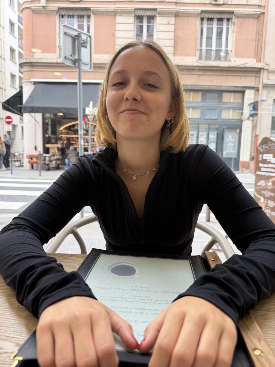 Elle assise en terrasse à Lyon, moue amusée, les mains posées sur un menu.