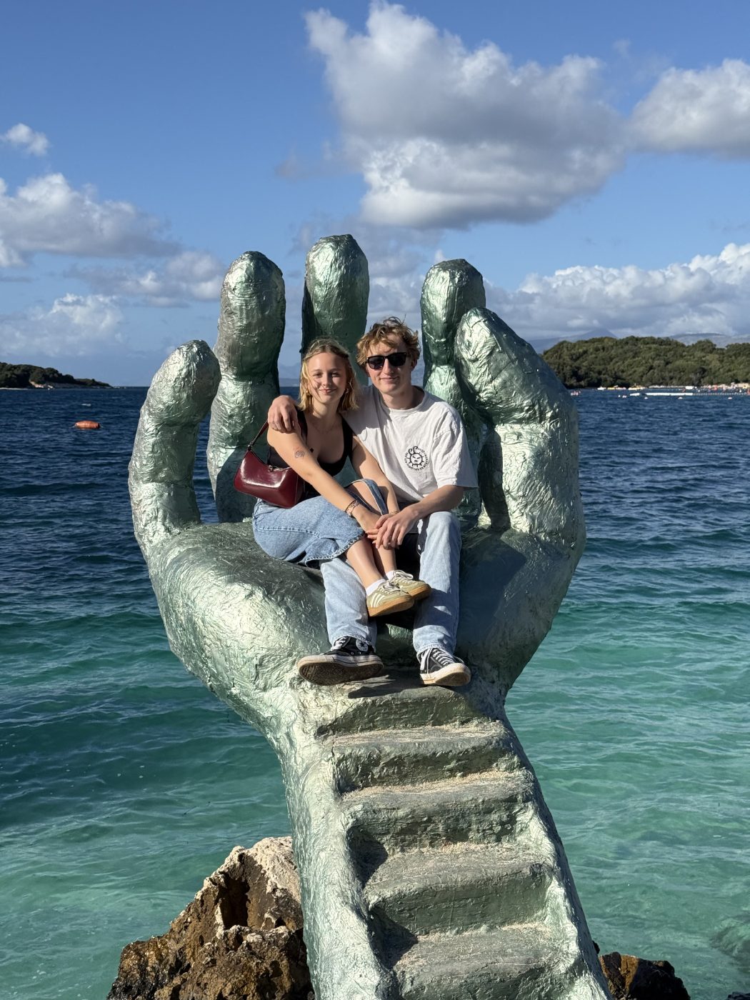 Tous les deux assis dans la paume d'une sculpture géante en forme de main, au-dessus d'une mer turquoise.