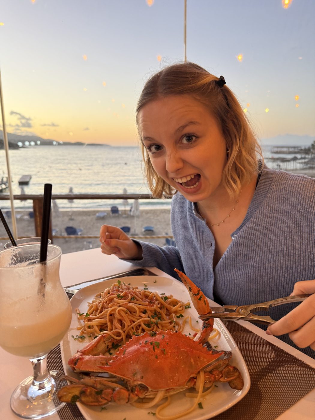 Au restaurant face à la plage au coucher du soleil : un crabe entier et des spaghettis, elle grimace de plaisir avec une pince en main.