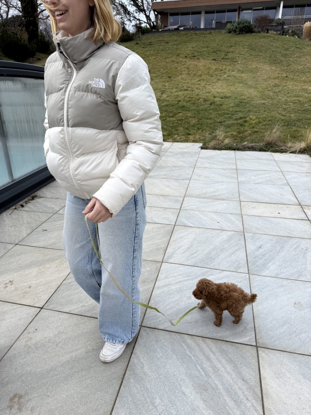 Sur une terrasse en hiver, elle en doudoune claire tient une laisse verte au bout de laquelle marche le chiot.