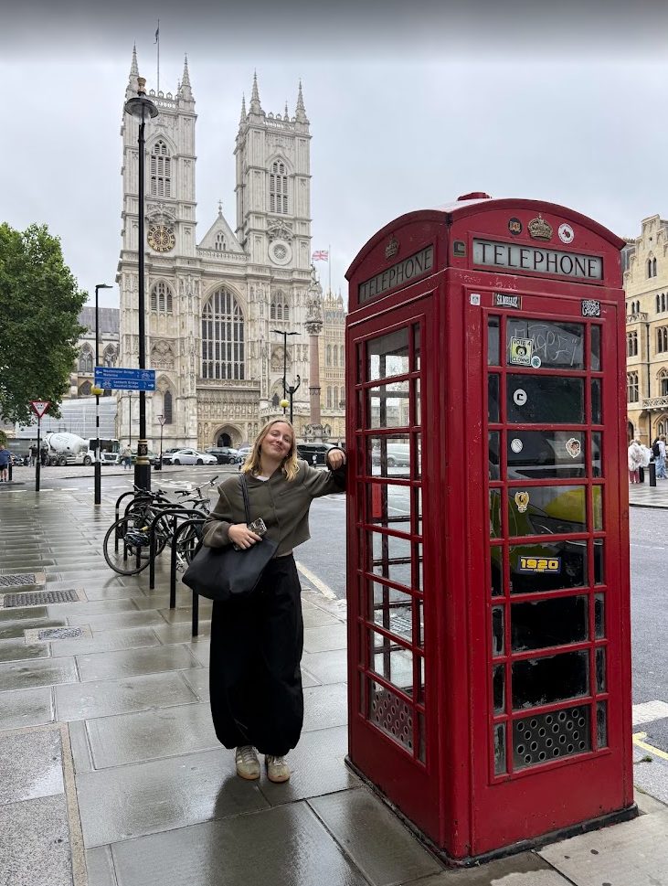 Elle appuyée contre une cabine téléphonique rouge sous un ciel gris, l'abbaye de Westminster derrière.