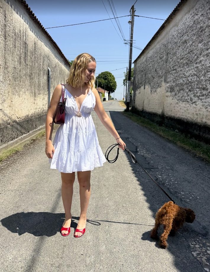 En plein soleil dans une ruelle entre deux murs blancs, elle marche en robe blanche et mules rouges, Ava au bout de la laisse.