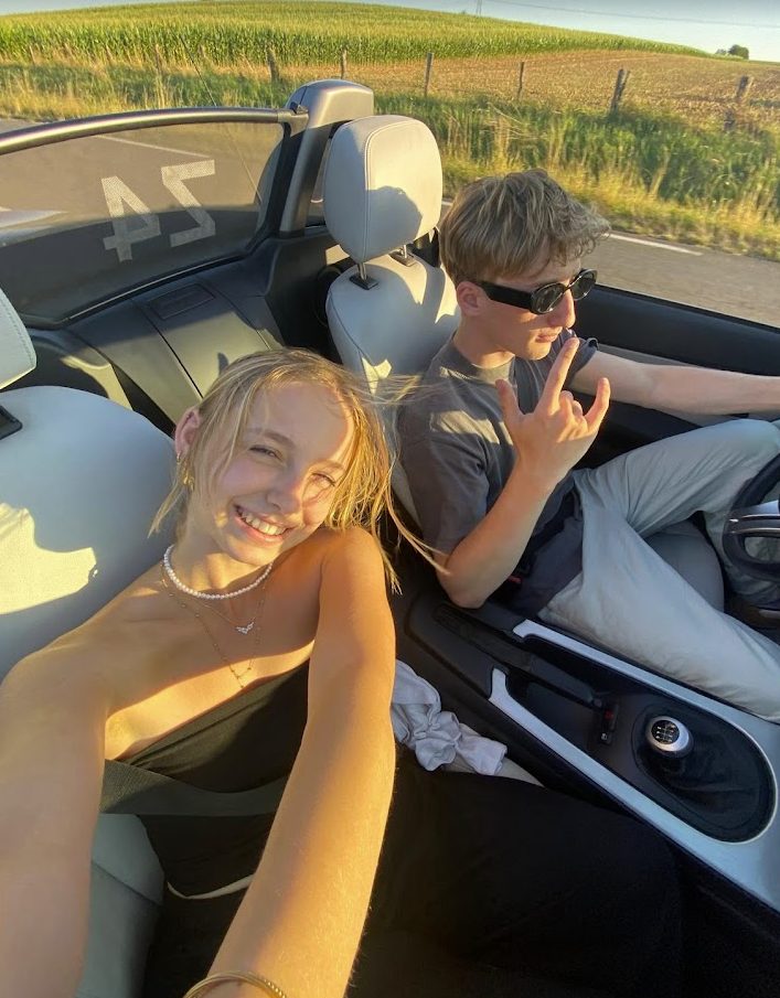 Selfie en décapotable au soleil rasant : elle rit à l'avant, lui conduit avec des lunettes noires, champs de maïs derrière.